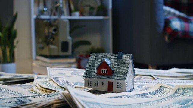 A tiny house sits atop a mound of cash in a cozy room. The scene highlights the relationship between money and home ownership, sparking ideas of investment and dreams