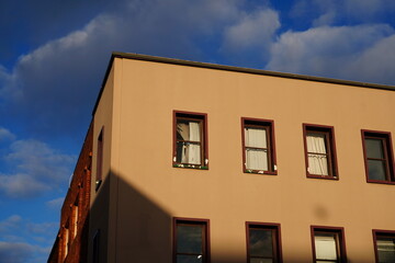 Obraz premium A medium shot of a vintage minimalist building facade with brown walls and reddish window frames, casting sharp geometric shadows under a cloudy blue sky.