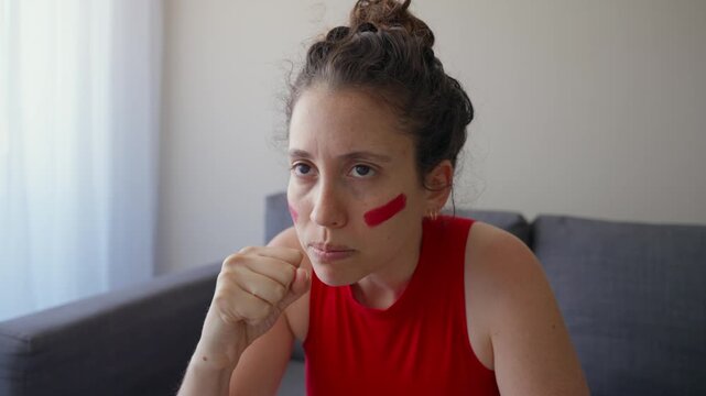 Passionate young woman watches game on TV excited but is disappointed by result. Female sports fan and supporter in red clothes and face paint for match stressed and anxious sad and angry with team