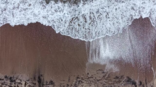Waves roll onto the shore of a sandy beach during sunset. The water creates foam as it meets the sand. The scene captures the beauty of nature at dusk.