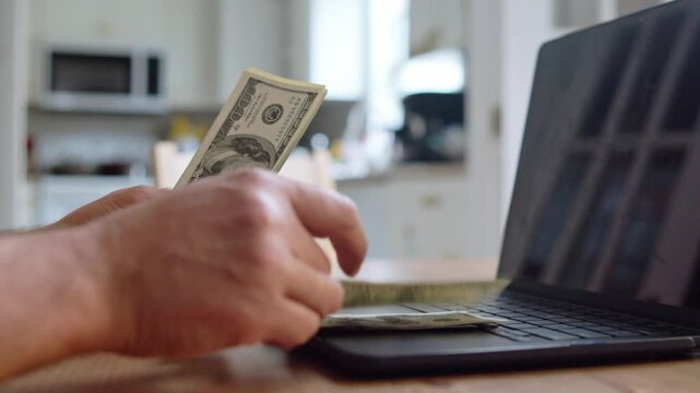 A hand counts crisp bills on a wooden table, focusing intently as numbers are crunched on a nearby laptop. An atmosphere of diligence fills the cozy kitchen