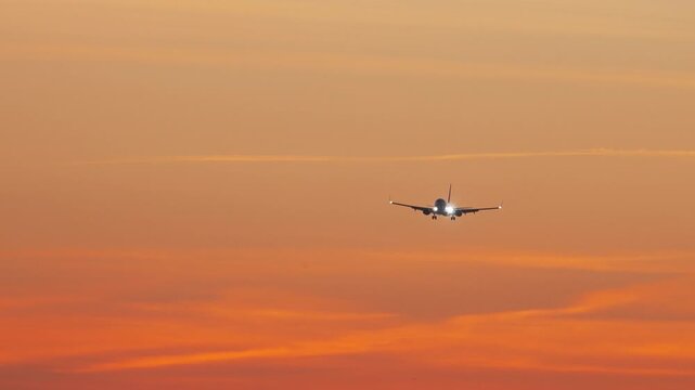 Commercial passenger jet on final approach with landing lights shining, descending against a vivid orange sunset sky streaked with cloud, an airliner arriving in the evening