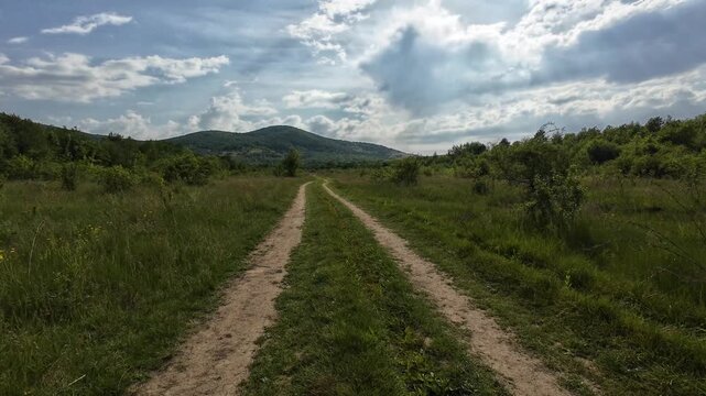 Point of view shot from a vehicle driving down a bumpy dirt track through a green valley toward a distant mountain under a dramatic cloudy sky during a bright summer day