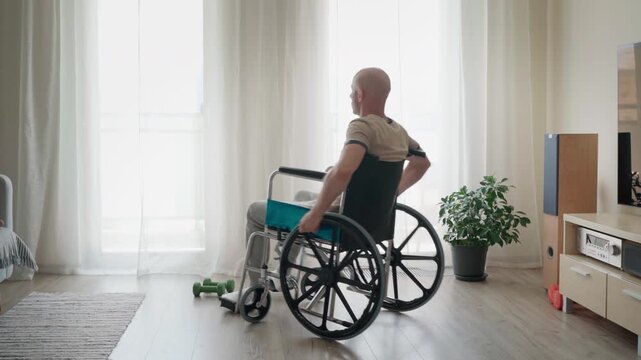 Sad bald man in a wheelchair sitting in a bright living room, rolling the wheels back and forth. Concept of loneliness, depression, physical disability, injury recovery, and boredom 4K video