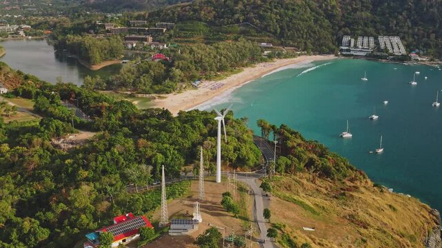 Wind turbine generating clean sustainable energy on a Phuket island cliff, tropical beach with yachts sailing in turquoise ocean. Showcasing environmental sustainability and tourism. Aerial panorama