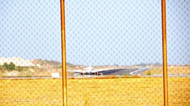 Airplane taxis past a chain-link fence. Yellow field stretches under clear skies. View framed by rusted gate posts. Scene suggests restricted airport access. Fence hides movement behind open sky