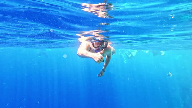 Underwater swimmer glides through calm, sunlit blue waters. Bubbles rise gently around the diver's movements. Single striped fish swims nearby, unbothered by human presence