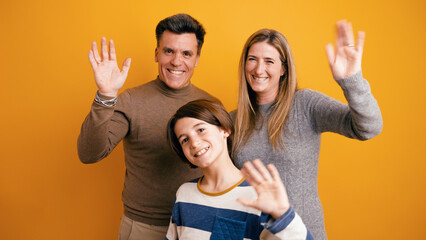 Obraz premium Joyful family with mother, father, and son waving hands, smiling with warmth, expressing a friendly greeting on a yellow background