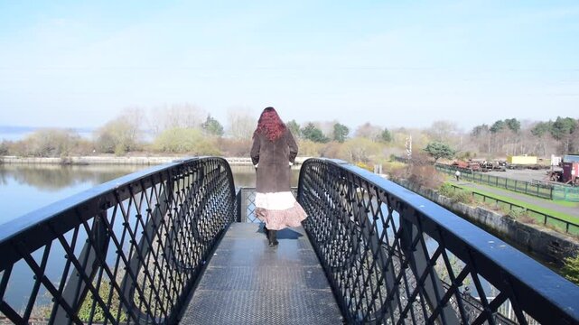 Close shot of a woman walking across a railway bridge on a bright spring day with trees and a river in the background