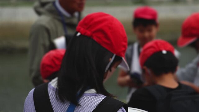 Group of school children students outdoor excursion, guided tour in the city with guide, docent with tourist pupils visitors, wear color caps hats, kids class field trip, urban sightseeing in Japan