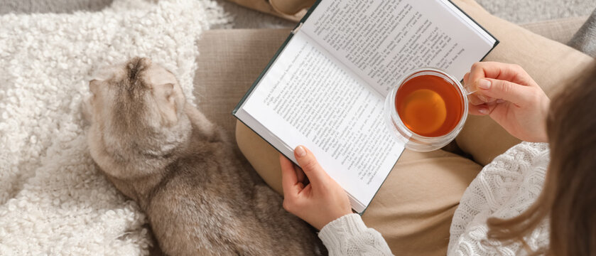 Young woman with cute cat reading book and drinking tea at home, top view