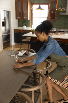 Long wooden dining table being set with glassware, woven placemats, plate, cutlery near window