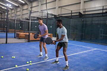 Fototapeta premium Diverse male friends holding padel rackets and practicing on blue court with scattered yellow balls