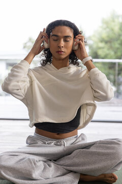 Female meditating on yoga mat by glass door holding headphones, wearing hoodie and smartwatch