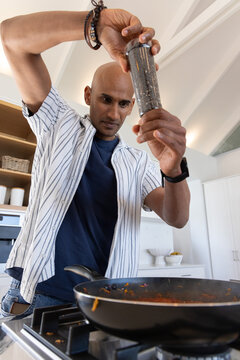 Indian man in 30s seasoning tomato sauce with pepper grinder over frying pan on gas cooktop
