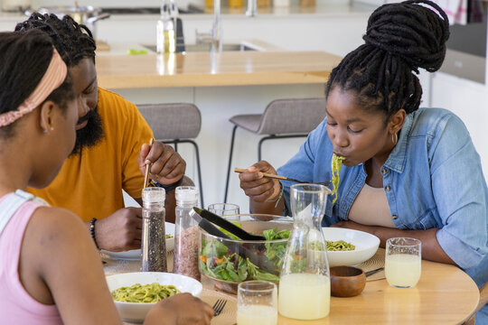 African American family sharing green pasta at wooden dining table in kitchen with salad bowl