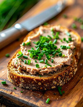 Fresh slice of multigrain bread with savory meat pate and chopped chives, metal knife on rustic wooden board, appetizing gourmet snack, tasty food. Close-up.