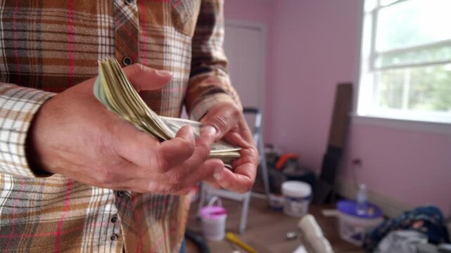 In a cozy room filled with renovation tools, a man carefully counts dollar bills to determine how much he will spend on materials for his home repairs