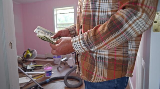 A man counts dollar bills in a room filled with renovation supplies, assessing the budget for materials needed to restore the space. His focused expression shows determination