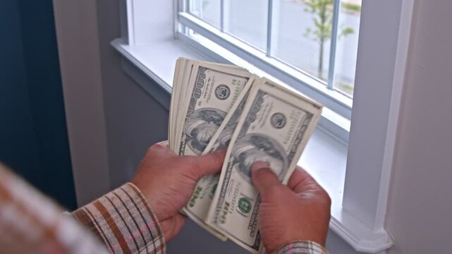 A man counts dollar bills, carefully estimating how much he needs for materials to repair his home. The bright room adds warmth to his thoughtful task