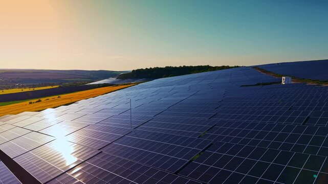 Rows of solar panels are installed on a hillside during sunset. The sun reflects off the panels. The landscape includes fields and trees in the background.
