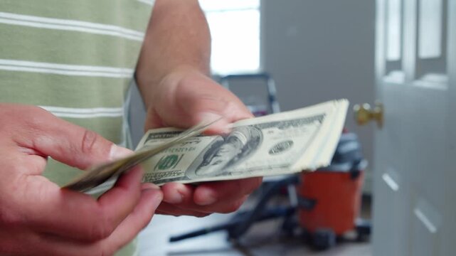 A man carefully counts his dollar bills to plan out expenses for necessary home repairs. The bright room is equipped with construction tools, creating a practical atmosphere
