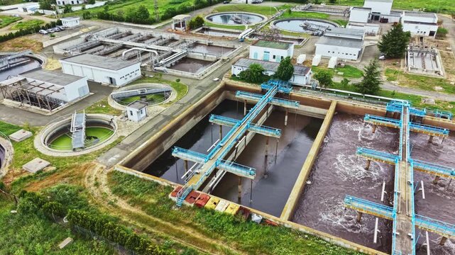 Aerial wastewater treatment plant aeration tanks, drone footage showing blue pipelines and mechanical aerators churning dark sludge, rectangular clarifiers