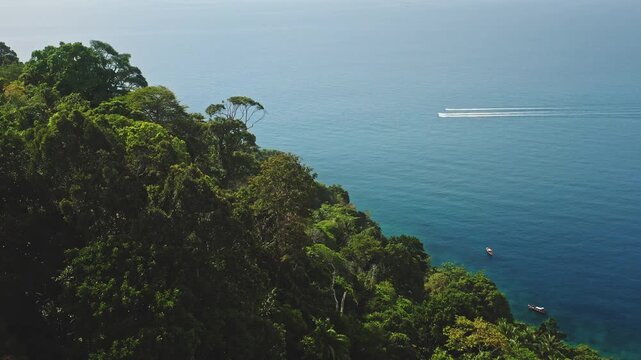 Phuket coastline where dense tropical jungle forest meets turquoise Andaman Sea, speedboats carving white wake while traditional longtail boats rest near sandy shore. Aerial view drone shot panorama