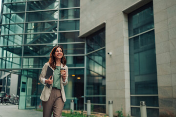 Businesswoman walking on urban street, carrying a laptop and coffee, smiling confidently while...