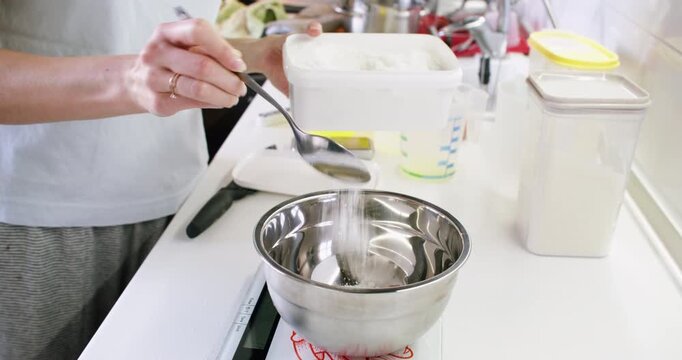 Kitchen workflow. Adding sugar. Female baker pouring sweet ingredient to metal bowl with spoon measuring recipe on table digital scale.