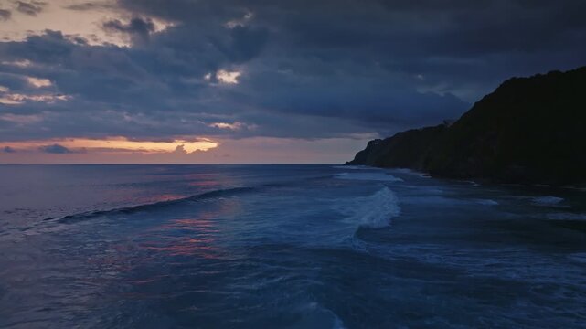 Ocean waves breaking on the shore of Bukit Peninsula are reflecting the colors of a dramatic sunset sky, creating a serene and captivating twilight scene. Blue toning night filter. Aerial drone flight