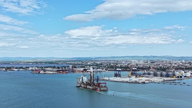 An industrial dredging vessel works in the port area. Nearby, various cargo ships and storage containers are visible. The sky is clear with some clouds in the background.