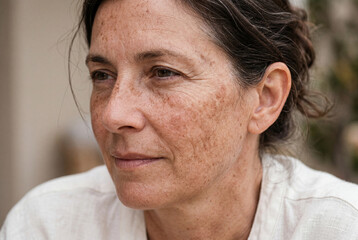 Naklejka premium A woman sits outside in daylight with a calm look on her face. She has freckles and brown hair tied back. The background shows a light, natural setting, creating an inviting mood