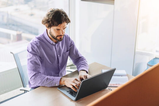 A man sits at a wooden desk and types on a laptop in a bright office. The city skyline can be seen through large windows. Papers are placed beside him on the desk
