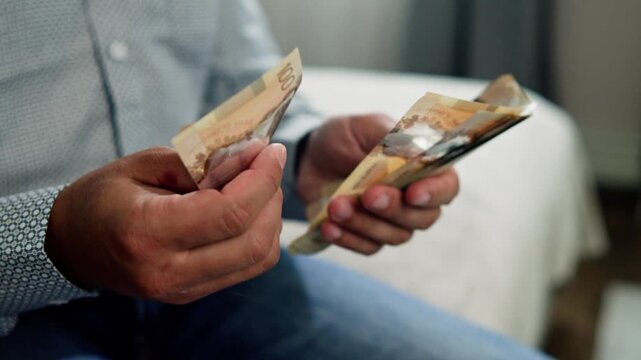 A man carefully counts Canadian dollar bills, his hands gently handling the money in a cozy room filled with a soft and inviting atmosphere, capturing a moment of focus and intent