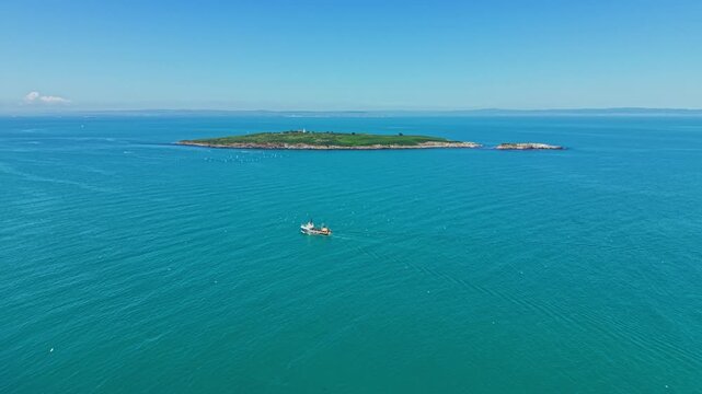 A boat travels across clear blue water near a small green island. The sky is clear with a few clouds visible in the distance. This scene shows a moment of nature and activity.
