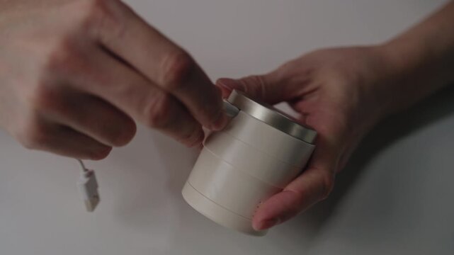 Top view of hands connecting white charging cable to portable electric lint remover machine at table, modern household appliance for clothing care and fabric maintenance. Shooting in slow motion.