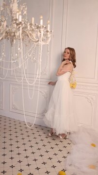 A young woman in a strapless white tulle wedding gown stands slightly left of center in a pale, ornate room with decorative wall paneling. She holds a sheer