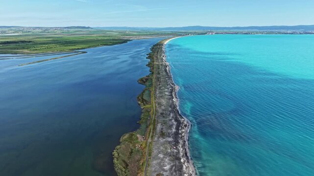 A long strip of land is seen separating a calm river from the ocean. The water is bright and blue. Green hills are visible in the distance under a clear sky. The scene shows a peaceful coastal area.