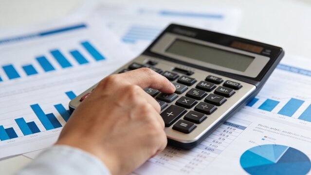 A professional financial analysis workspace, close-up of a hand pressing buttons on a modern calculator placed over printed business reports and analytics charts. Blue bar graphs, pie charts, and fina