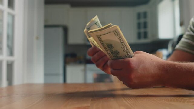 A man sits comfortably at a wooden table in his cozy home, counting stacks of dollar bills with focused attentiveness, enjoying a quiet moment of reflection
