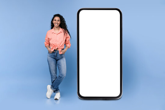 Woman with curly hair wears a pink shirt and blue jeans. She stands next to a large blank smartphone on a blue background. The setting has bright lighting and a clean appearance