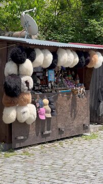 Traditional souvenir shop on a cobblestone street in beautiful Georgia, displaying fluffy sheepskin papakha hats, local crafts, magnets, and baseball caps for tourists on a sunny summer day.

