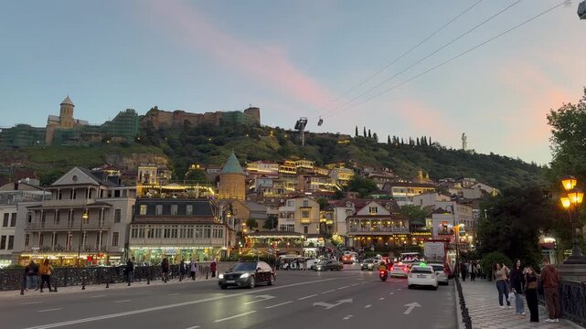 Scenic evening view of Tbilisi old town in Georgia, featuring the illuminated Narikala fortress on the hill, Metekhi church, passing cable cars, and busy street traffic at sunset.

