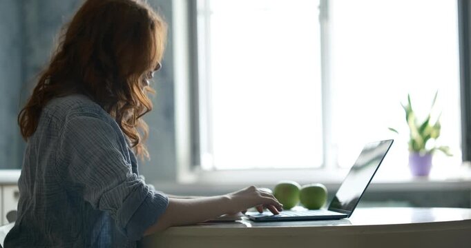 teenage girl with vibrant red hair sitting at a kitchen counter, focused on her laptop computer. She is actively typing and looking intently at the screen, writing or working on a digital project.