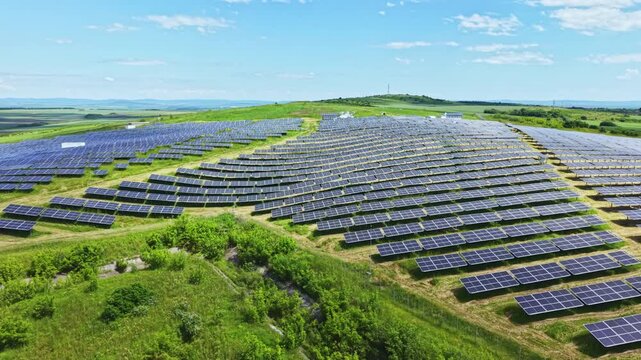 Aerial solar panel field sweeping rows across rolling green hills under clear blue sky shimmering photovoltaic modules aligned in grid patterns, drone survey