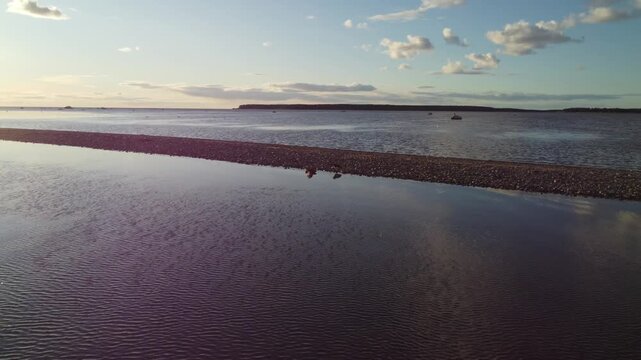 Aerial circular orbit view of a flock of seabirds resting and nesting on a narrow gravel sandspit near Pandju island bird sanctuary in Viimsi, Estonia, surrounded by calm sea water during sunset.