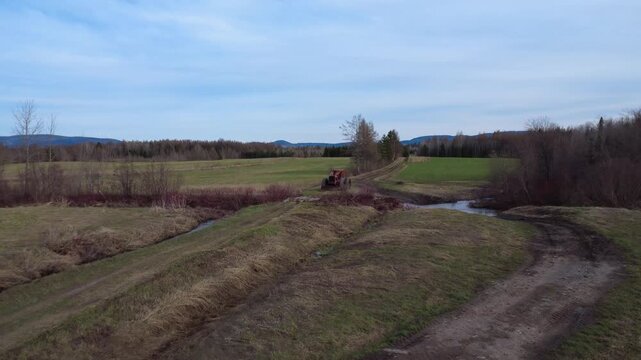Old red Timberjack skidder parked on a muddy dirt road beside a rustic log bridge, overlooking green fields, streams and forest during early spring.