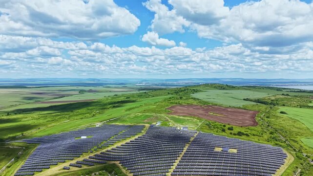Aerial solar farm on rolling hills, spiral arrays of photovoltaic panels carving patterns into vibrant green landscape under wide cloudscape, distant horizon
