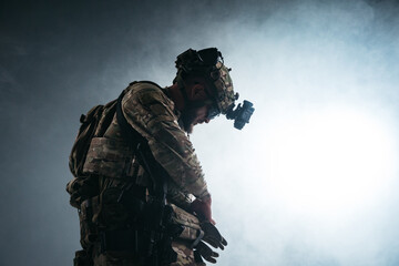 Profile portrait of a tactical soldier in camouflage uniform with night vision helmet standing in smoky studio lighting, representing military readiness, defense concept. Free copy space © Валерия Стоганенко